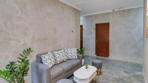 Ein modernes Wohnzimmer mit einem grauen Sofa, einem weißen Couchtisch und zwei grauen Hocker. Im Hintergrund sind Blumentöpfe und eine Holztür zu sehen.