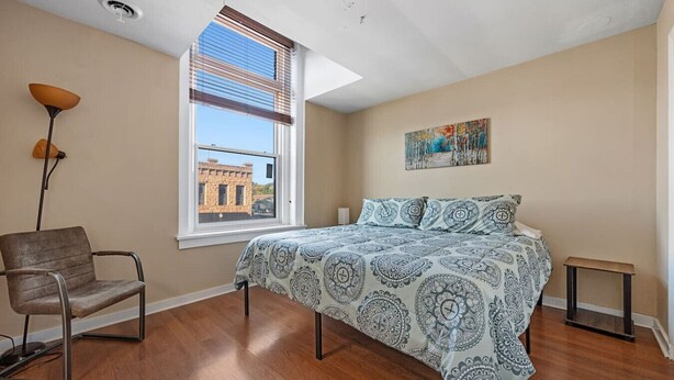 Una camera da letto con un letto, una sedia, un tavolino, una finestra con persiane e un quadro appeso al muro.