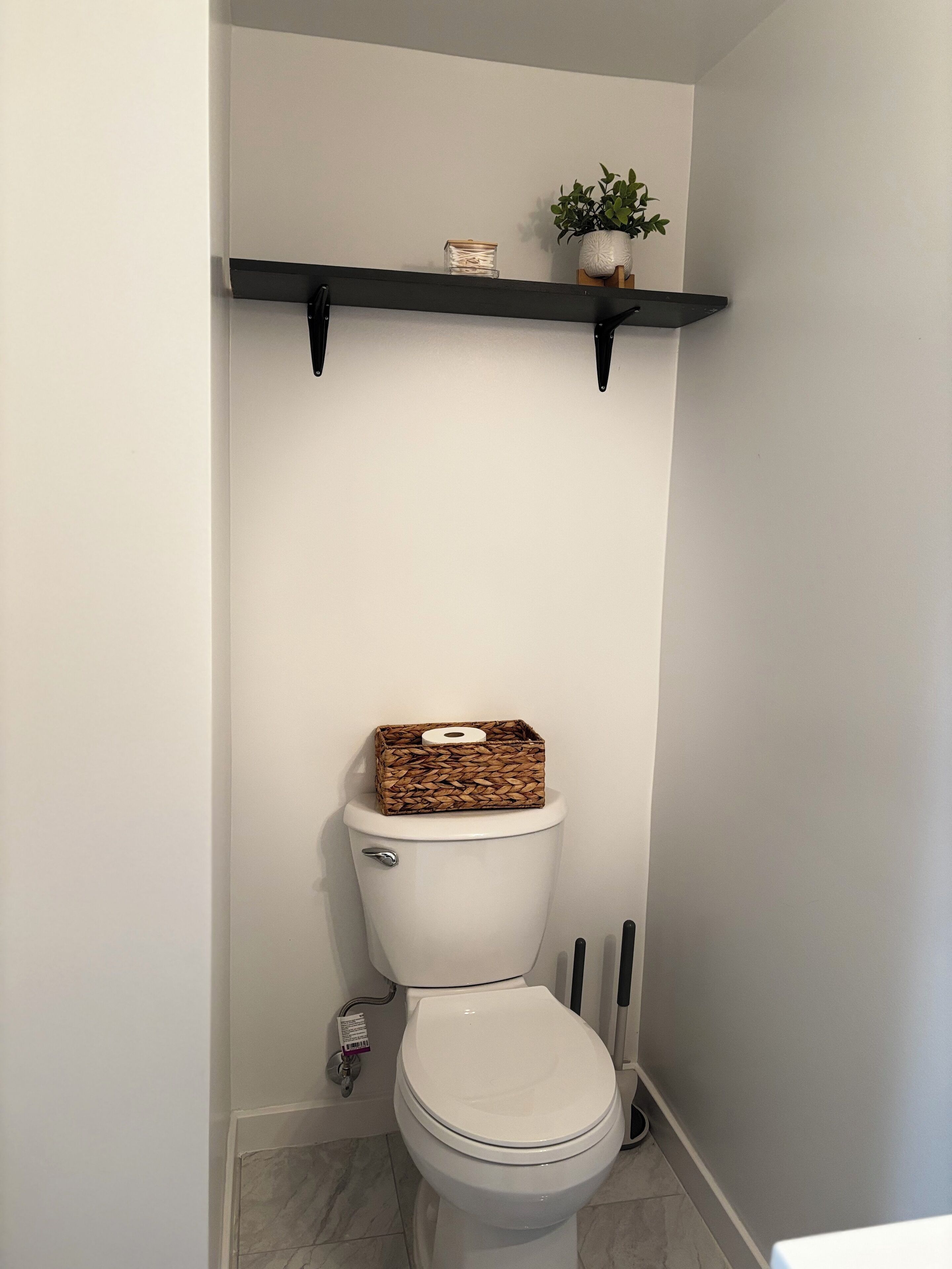 Des toilettes avec un panier et un rouleau de papier toilette posés sur le dessus, une étagère au-dessus avec une plante et une bougie, et un sol en marbre.