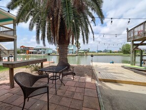 Een terras met rieten meubilair, een palmboom en uitzicht op een waterpartij met gebouwen op de achtergrond.