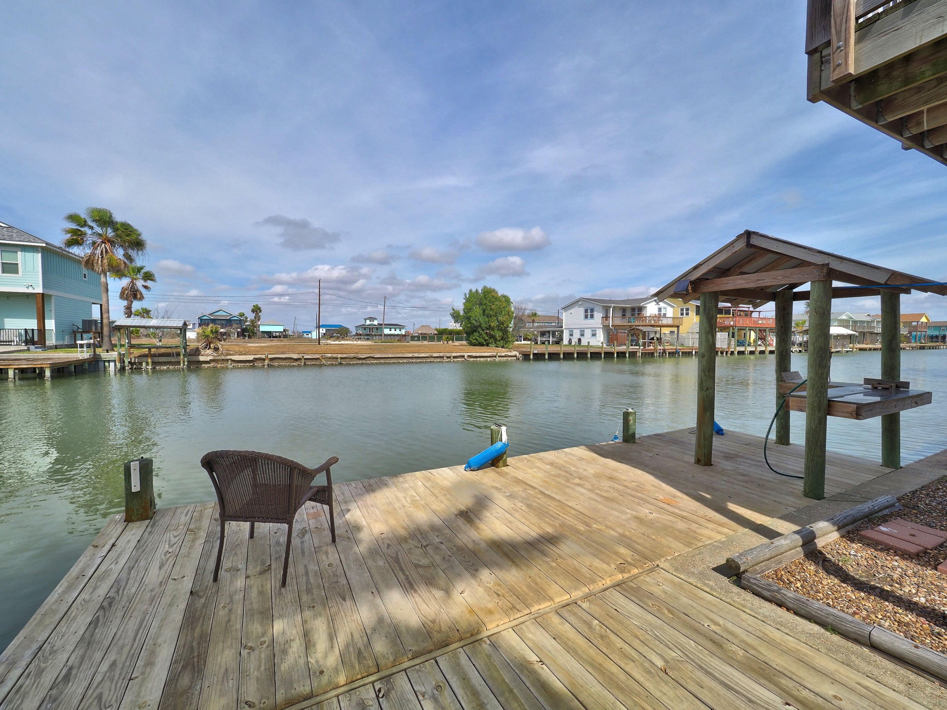 Un muelle de madera con una silla, un área techada y vistas a casas al otro lado de un cuerpo de agua tranquilo.