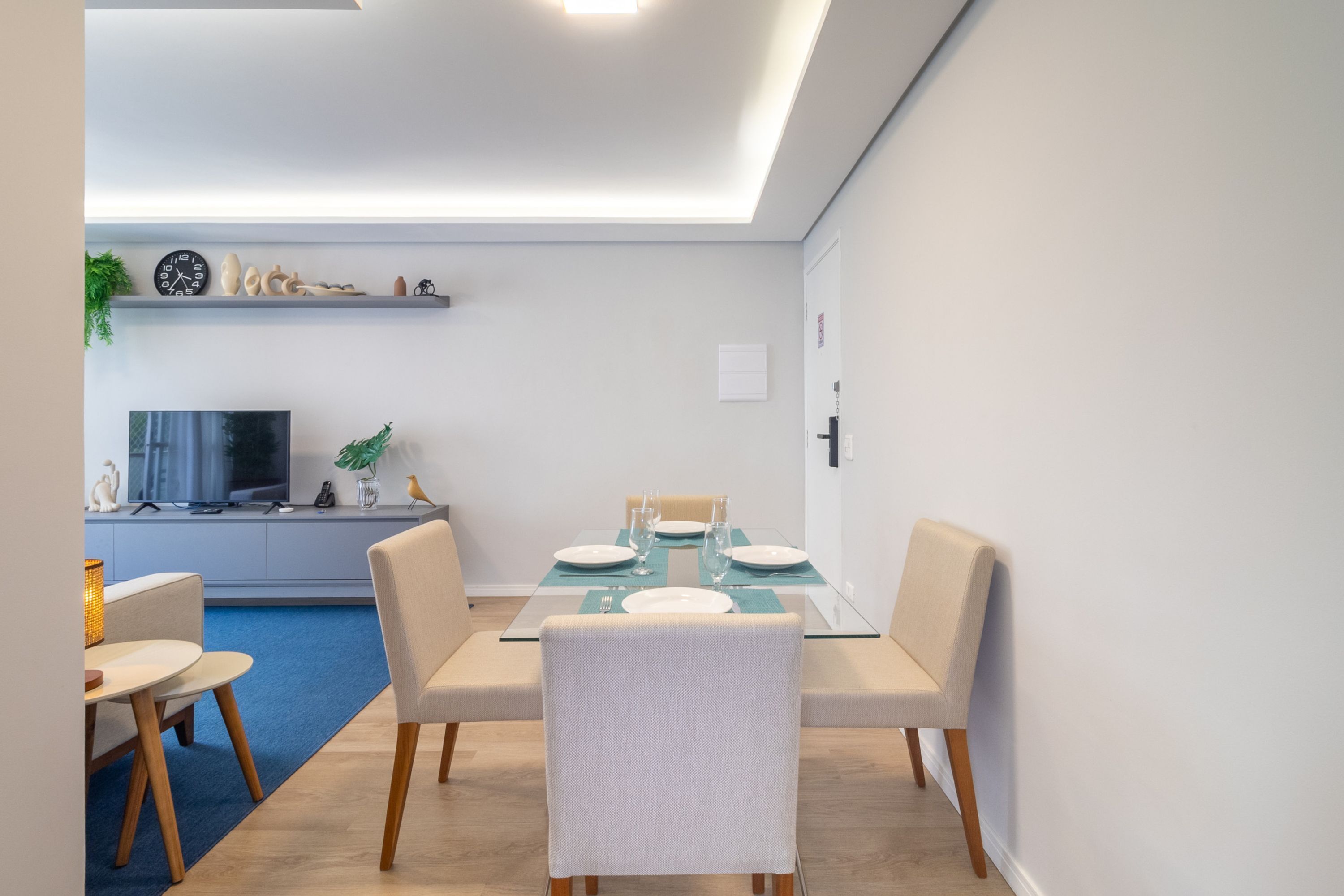 A modern dining area with a glass table, white chairs, a flat-screen TV, and a blue carpet.