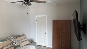 Una stanza con una ventola a soffitto, una porta bianca, un armadio in legno, un divano grigio con cuscini decorati e una televisione a schermo piatto montata al muro.