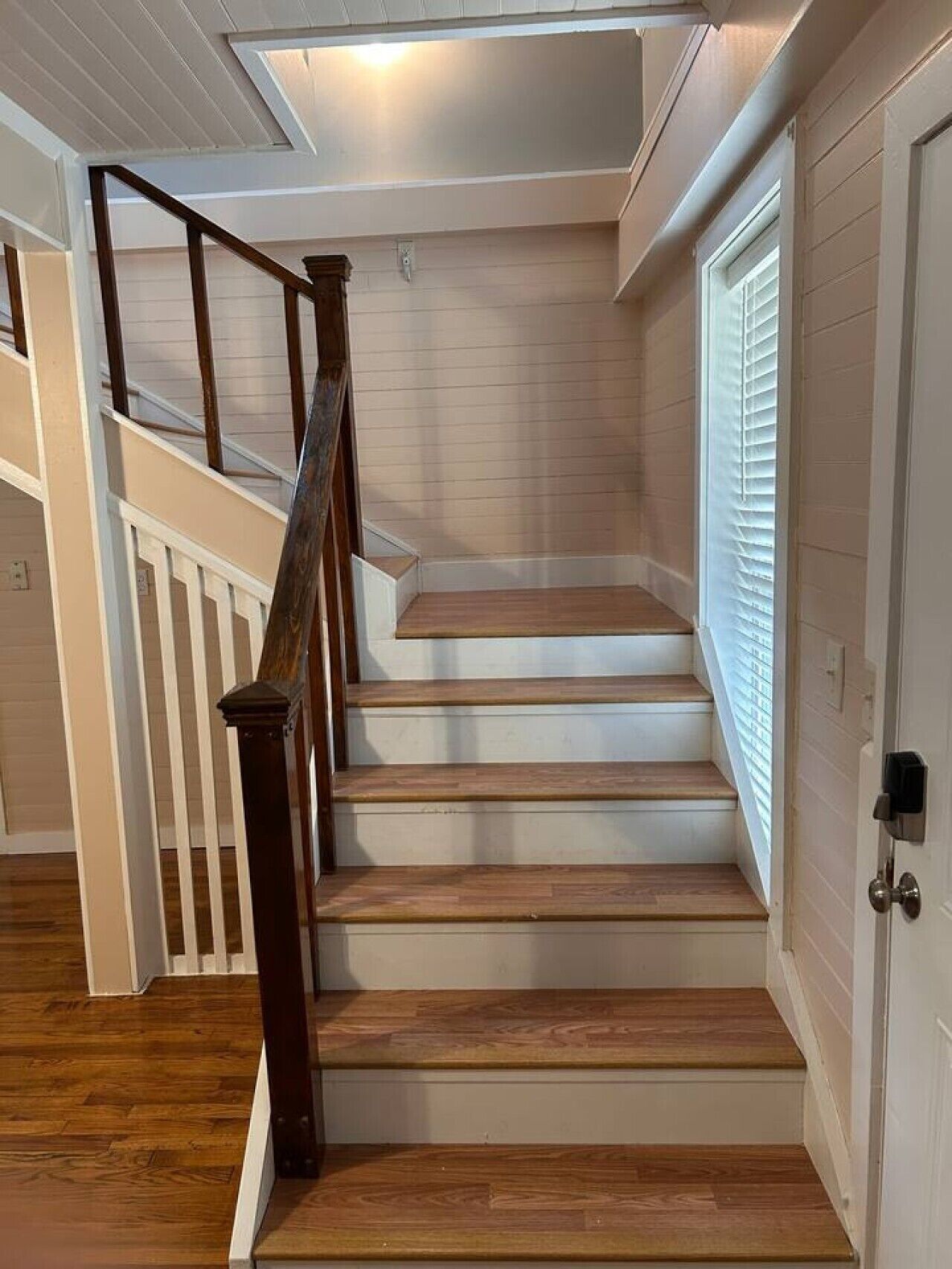 A staircase with wooden steps and a wooden handrail, leading to a landing with a white door and a window with blinds.