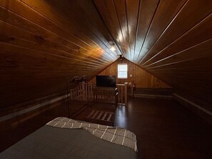 Ein Zimmer mit Holzwänden und einer geneigten Decke, ein Bett mit einem gemusterten Überwurf, ein Fernseher auf einem Beistelltisch und ein kleines Fenster, das für natürliches Licht sorgt.