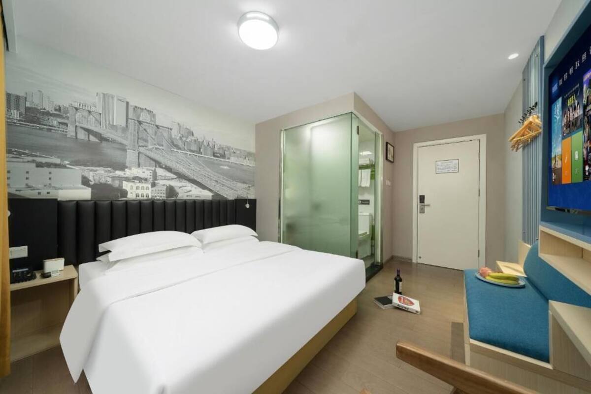 A hotel room with a bed, a cityscape mural, a glass door closet, and a small seating area with a table.