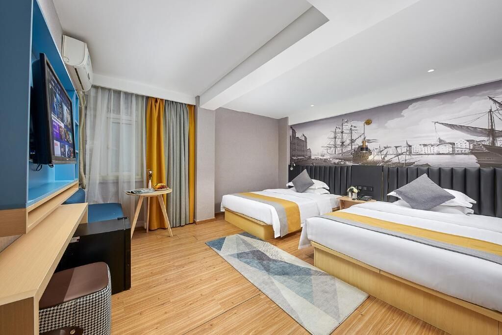 A hotel room with two beds, a desk, a television, and a mural of ships on the wall.