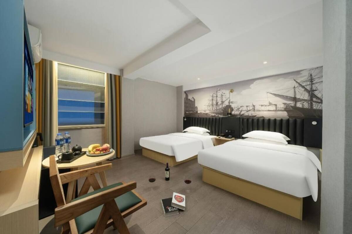 A hotel room with two beds, a desk with a chair, a small table with food and drinks, and a large window.