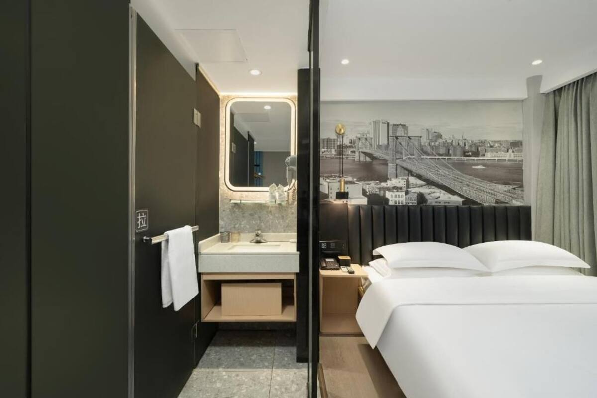 A modern hotel room with a bed, a bathroom with a mirror, and a cityscape view.