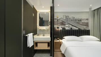 A modern hotel room with a bed, a bathroom with a mirror, and a cityscape view.