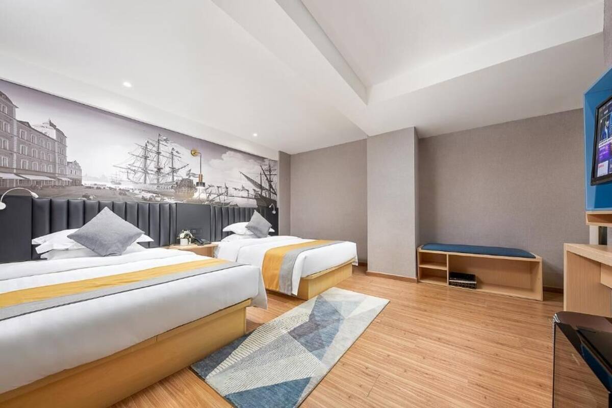 A hotel room with two beds, a large mural, a desk, and a bench.