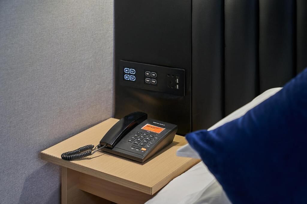 A hotel room with a bedside telephone, a control panel with buttons, and a bed with a blue pillow.