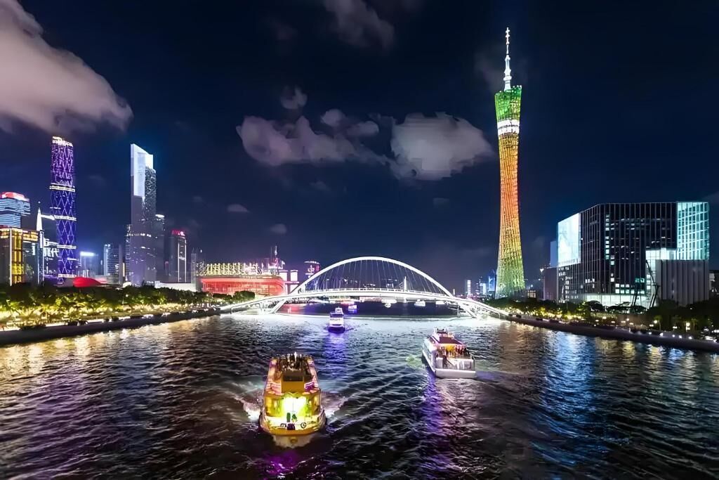 A nighttime cityscape with illuminated buildings, a bridge, and boats on a river.