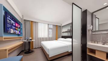 A hotel room with a bed, a bedside table, a TV mounted on the wall, a bathroom with a mirror and sink, and a cityscape view.