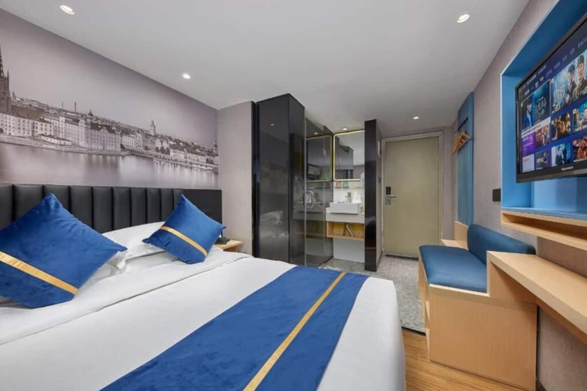 A hotel room with a bed, a desk, a chair, a cityscape mural, and a TV screen.