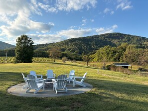 Eine Terrasse mit Adirondack-Stühlen und einer Feuerstelle aus Stein, umgeben von einer Wiese und einem bewaldeten Hügel.