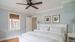 Un dormitorio con cama, ventilador de techo, ventana con persianas, puerta, mesita de noche y dos cuadros en la pared.