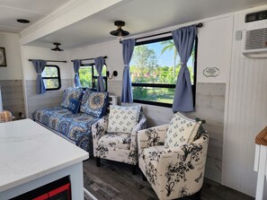 Een gezellig interieur van een camper, met een bed, twee stoelen, een tafel en grote ramen met gordijnen.