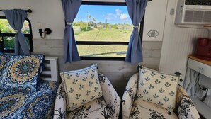 A cozy interior with a bed, a window with curtains, a wall-mounted light, and a small table with a red object. - 12 min to the Everglades Nat. Park & near FL Keys! Cozy RV in Redland w/laundry (Homestead)