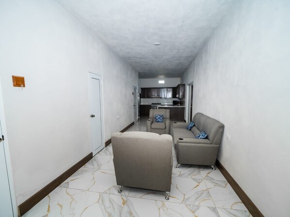 A narrow hallway with a door on the left, a sofa and armchair in the center, and a kitchen in the background.