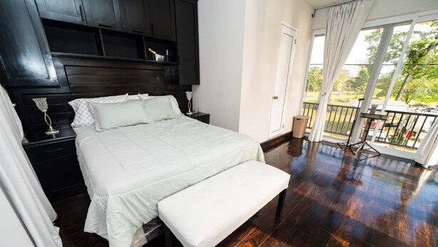 A bedroom with a large bed, a bedside table, a nightstand, a bench, a balcony with a railing, and a view of greenery outside.