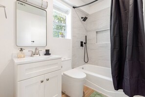A modern bathroom with a white vanity, a large mirror, a shower with a handheld showerhead, and a black shower curtain. - Renovated Brentwood Home Near DT, Domain & Q2 (Austin)