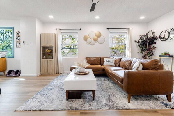A modern living room with a brown leather sofa, white coffee table, and a large window with curtains. - Renovated Brentwood Home Near DT, Domain & Q2 (Austin)