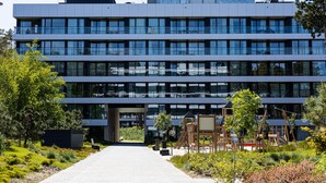 Un edificio moderno de varios pisos con una entrada central, rodeado de vegetación y juegos infantiles al aire libre.