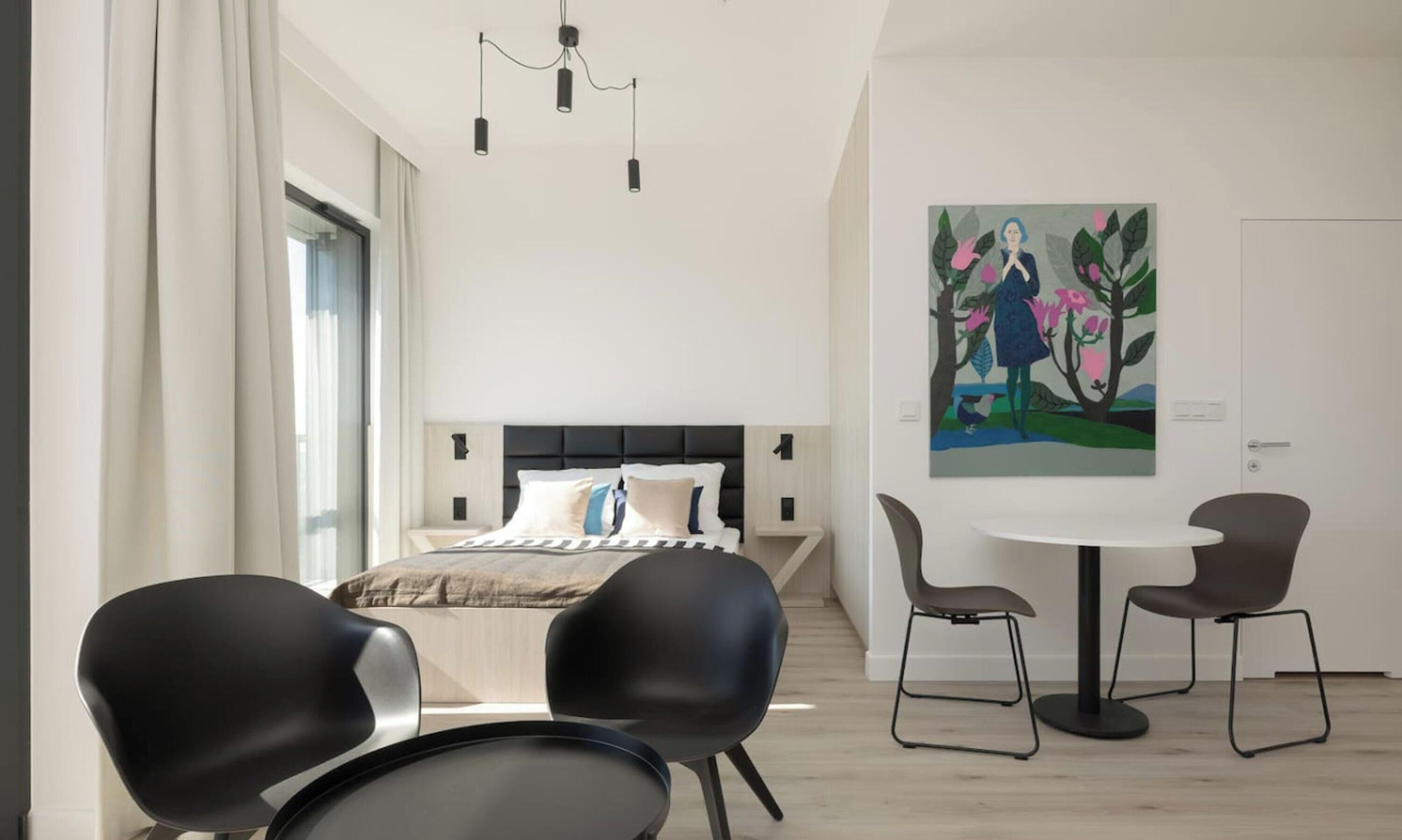A modern bedroom with a bed, two black chairs, a round table, and a painting on the wall.