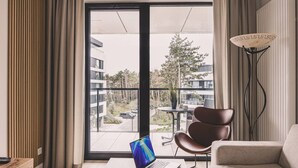 Moderni olohuone, jossa on pöydällä kannettava tietokone, sohva, nojatuoli, lattiavalaisin ja suuri ikkuna, josta avautuu näkymä parvekkeelle ja ympäröiville rakennuksille.