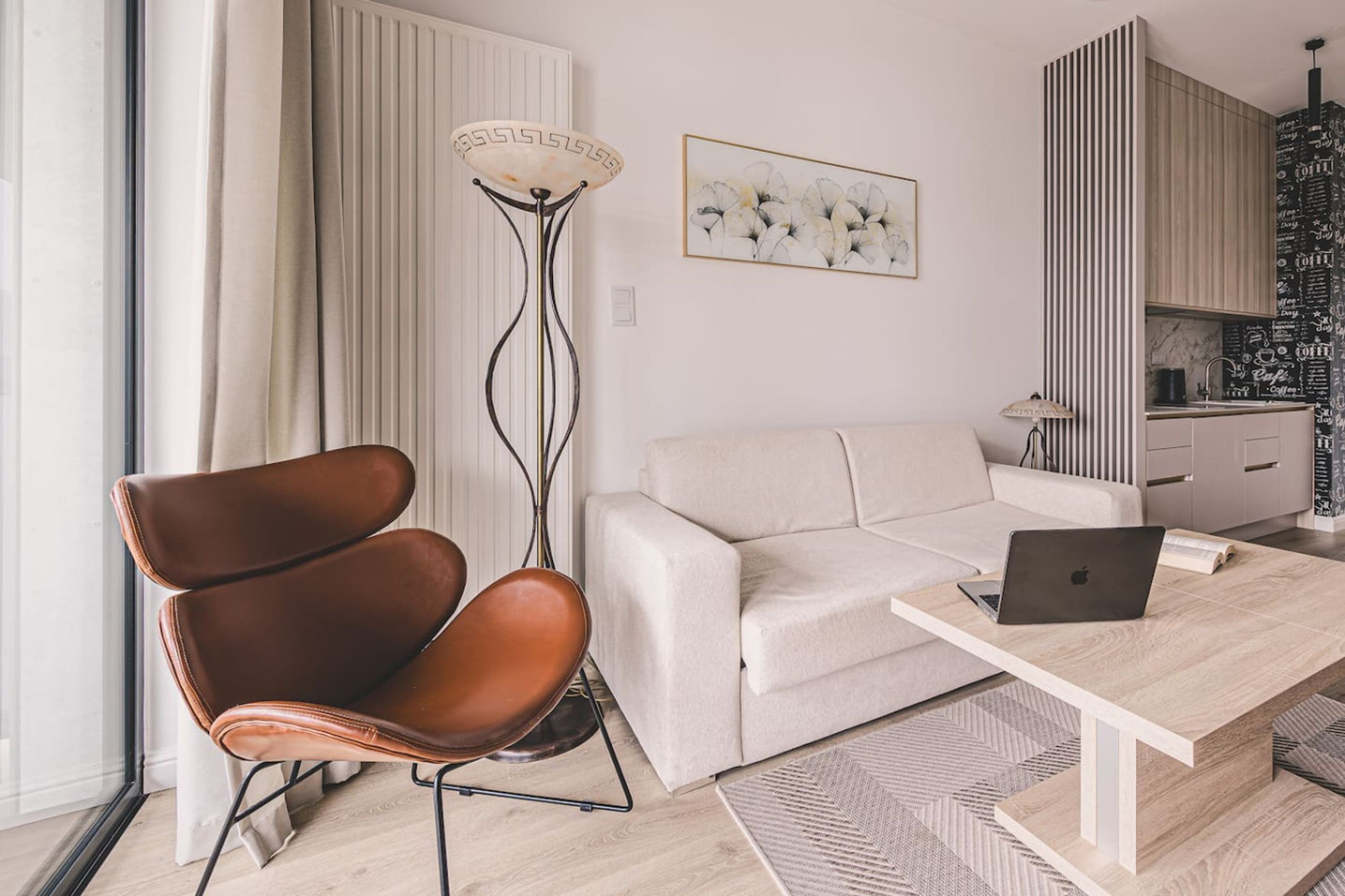 A modern living room with a sofa, a brown chair, a coffee table with a laptop, a floor lamp, and a kitchen area with a patterned backsplash.