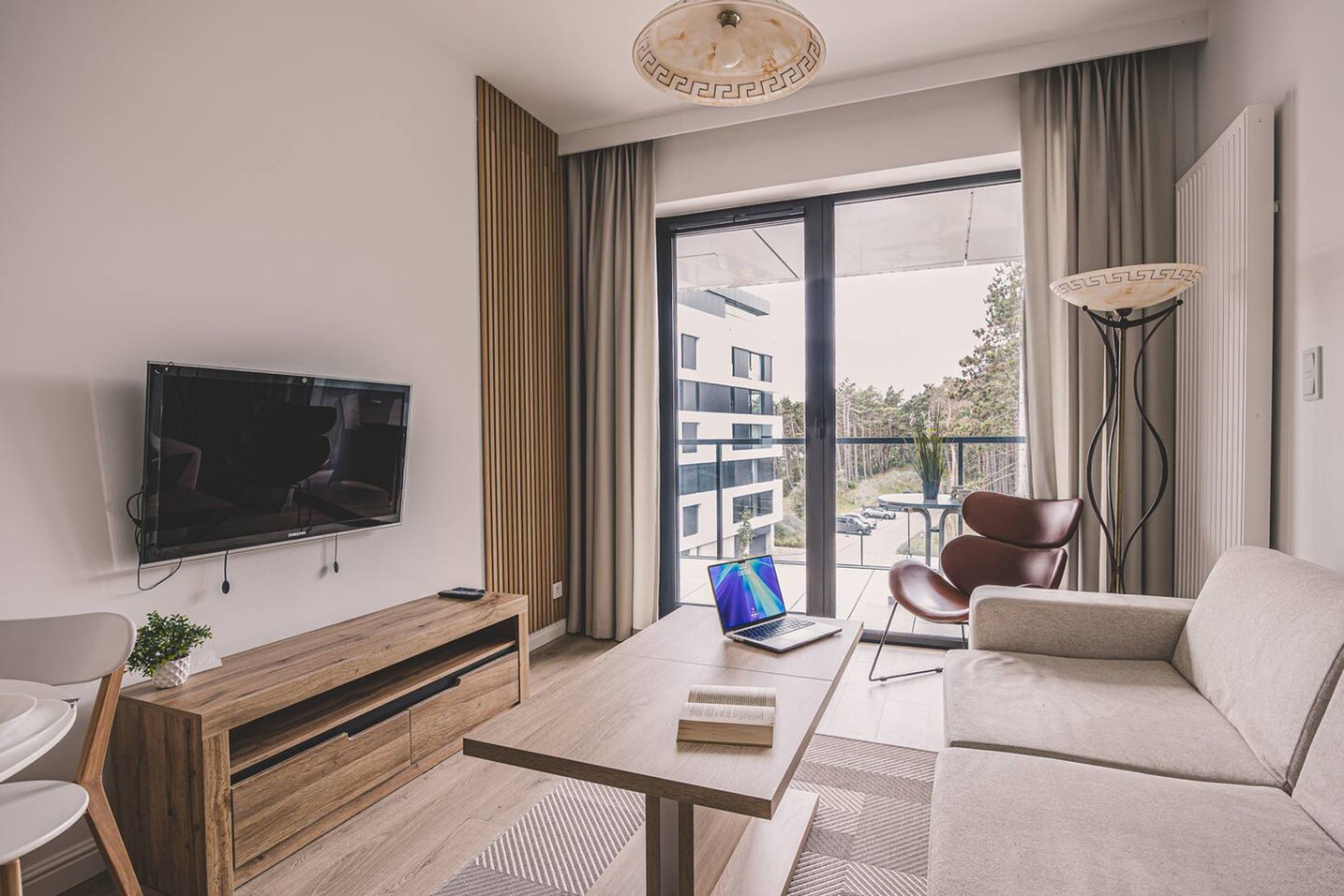 A modern living room with a flat-screen TV, a laptop, a sofa, a coffee table, and a view of buildings through a large window.