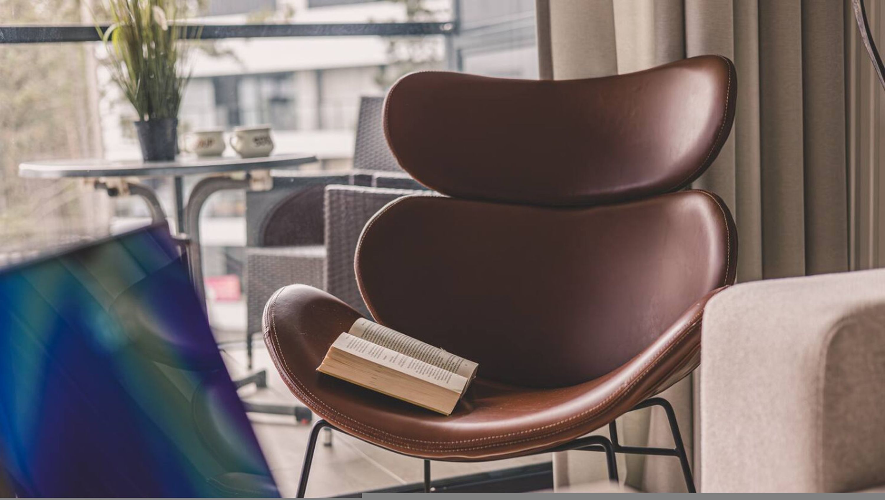 Una silla de cuero marrón con un libro, una mesita con una planta y dos tazas, y una ventana con cortinas.
