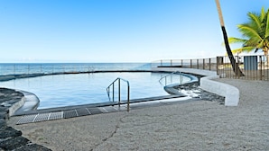 A circular infinity pool with a concrete edge, a metal safety railing, and a pebble beach surrounding it. - Casa De Emdeko 303 | Ocean View | Central AC (Kailua Kona)