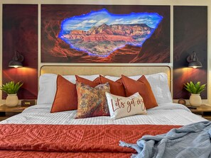 Un lit avec une tête de lit blanche, plusieurs oreillers, une couverture rouge et un téléviseur fixé au mur qui diffuse un paysage.