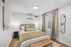 A modern bedroom with a bed, bedside lamps, a bedside table, a window with curtains, a wall-mounted air conditioner, and a mirror. - Large Clean & Modern Basement Apartment Near Aggie (Manhattan)