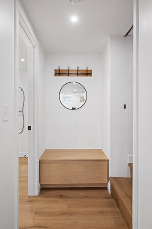 A narrow hallway with a wooden bench, a round mirror on the wall, and a wooden shelf above. - Large Clean & Modern Basement Apartment Near Aggie (Manhattan)