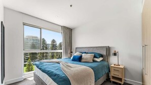 Ein modernes Schlafzimmer mit großem Fenster, einem Bett mit blauer Bettwäsche, einem Nachttisch und einem Kleiderschrank.