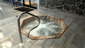 A hot tub with a wooden frame and a metal ladder, set on a tiled floor with a stone wall in the background. - Cozy 1-bedroom condo in BEECH MOUNTAIN/Hike/Bike/Pool/Raft/Tennis/Pickleball (Beech Mountain)