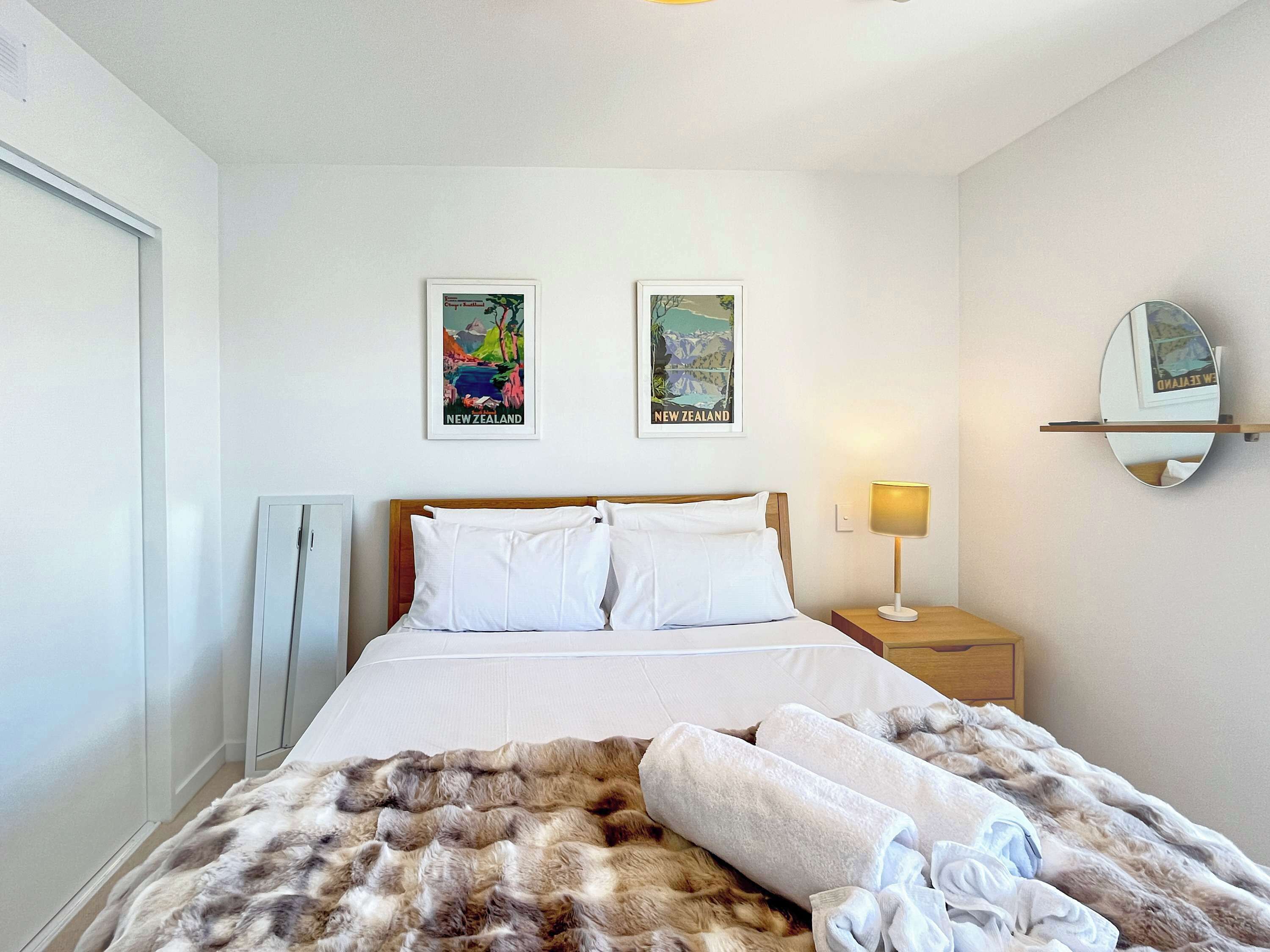 Habitación de hotel con cama, dos carteles enmarcados de viajes, una lámpara de noche y un espejo en la pared.