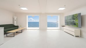 A modern living room with a green sofa, a wooden coffee table, a white TV on the wall, and a large window offering a view of the sea. - Daebudo Sunbell Ocean View Pension (Ansan)