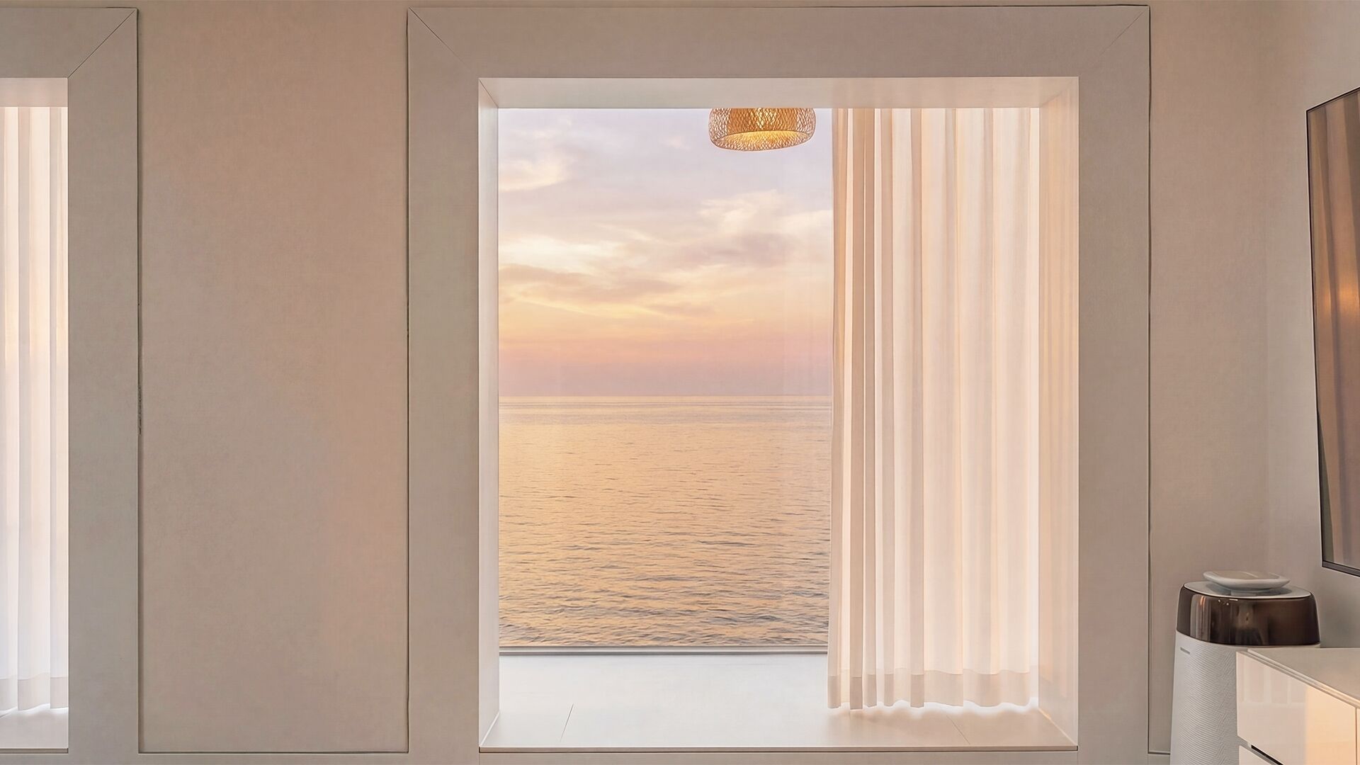A room with a view of the sea at sunset, a window with curtains, and a small table with a lamp.