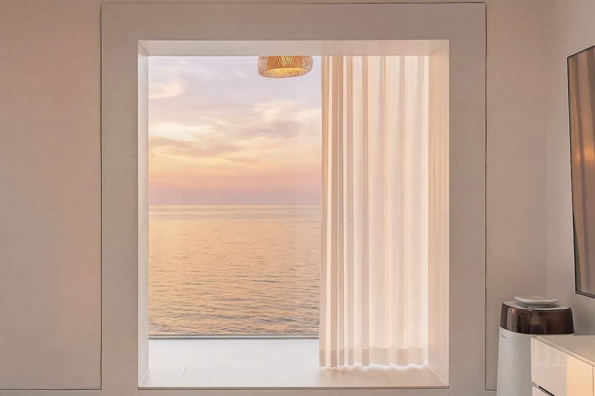 A room with a view of the sea at sunset, a window with curtains, and a small table with a lamp.
