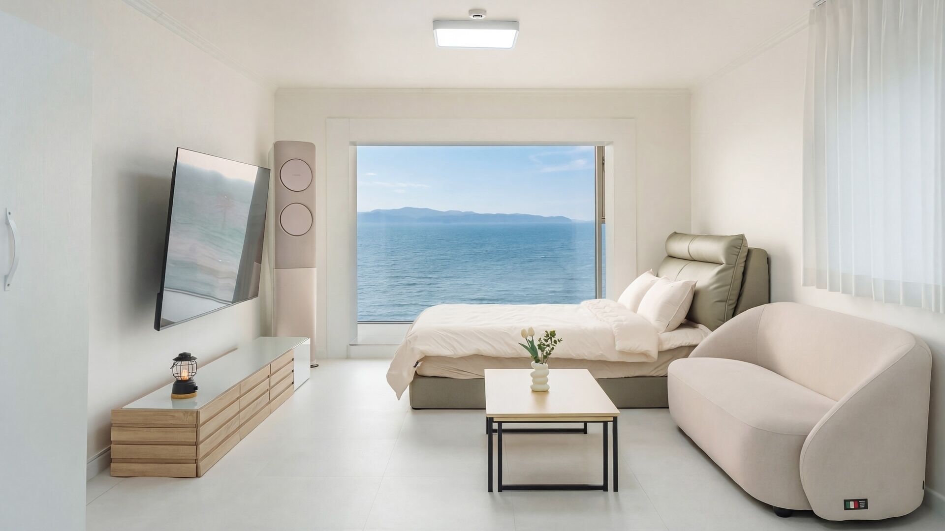 A modern bedroom with a bed, a sofa, a TV, a table, and a view of the sea.