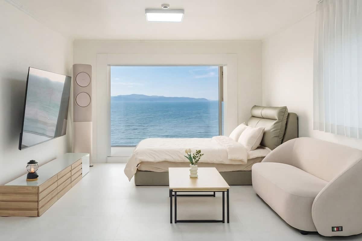 A modern bedroom with a bed, a sofa, a TV, a table, and a view of the sea.
