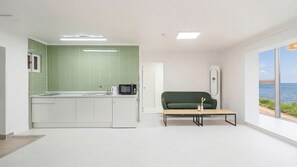 A modern kitchen with a green backsplash, a microwave, and a sink. There is a green sofa and a small table with a vase. A large window offers a view of the sea.