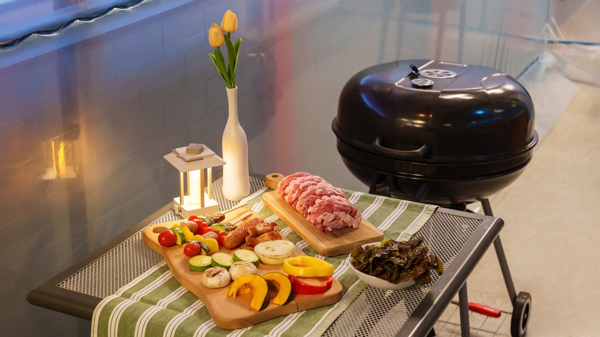 Uma churrasqueira portátil com carne crua e vegetais em uma mesa, um vaso com flores e um pequeno candeeiro.