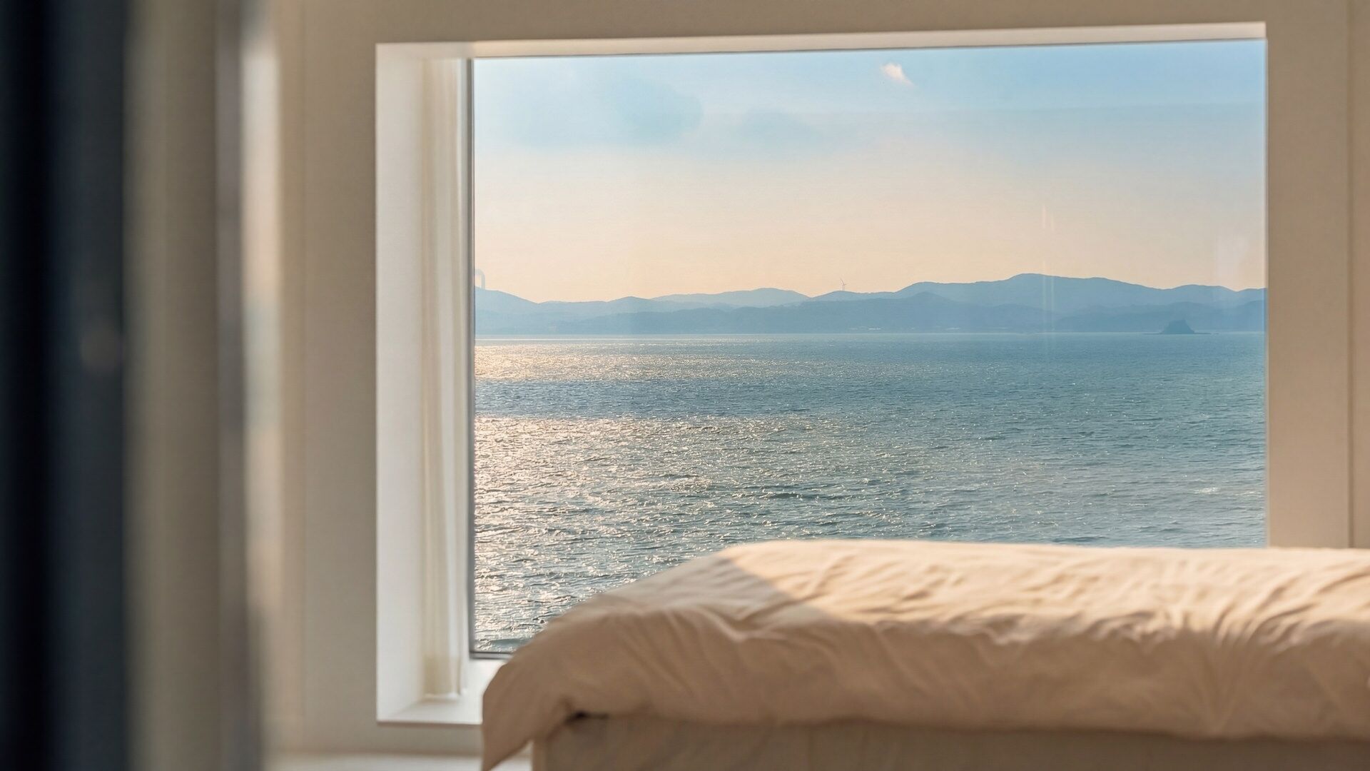 A bed with a white duvet, a window with a view of a calm sea and distant mountains.