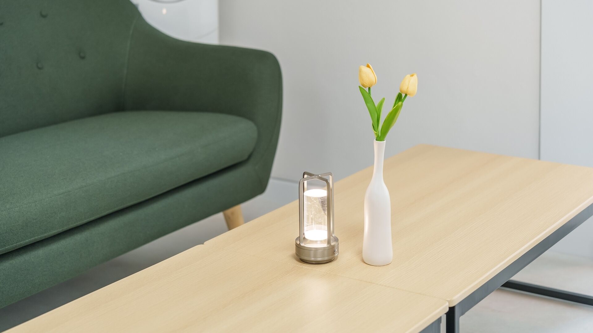 A modern room with a green sofa, a wooden table with a white vase and yellow flowers, and a lit candle.