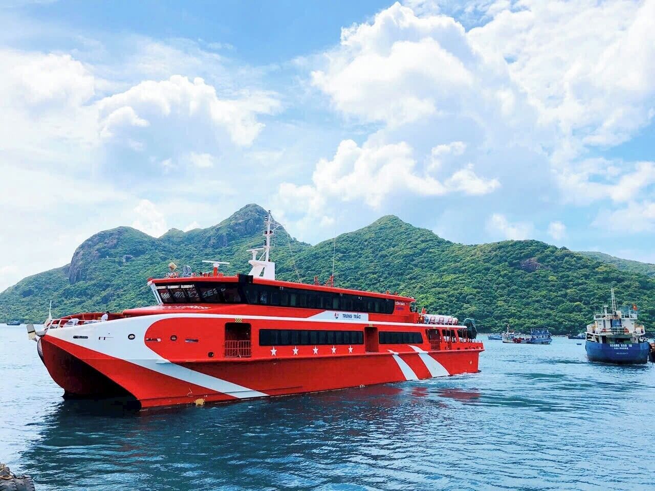 Een rode en witte boot met de tekst 'TRUNG TRANG' erop, afgemeerd in rustig water, met bergen op de achtergrond.